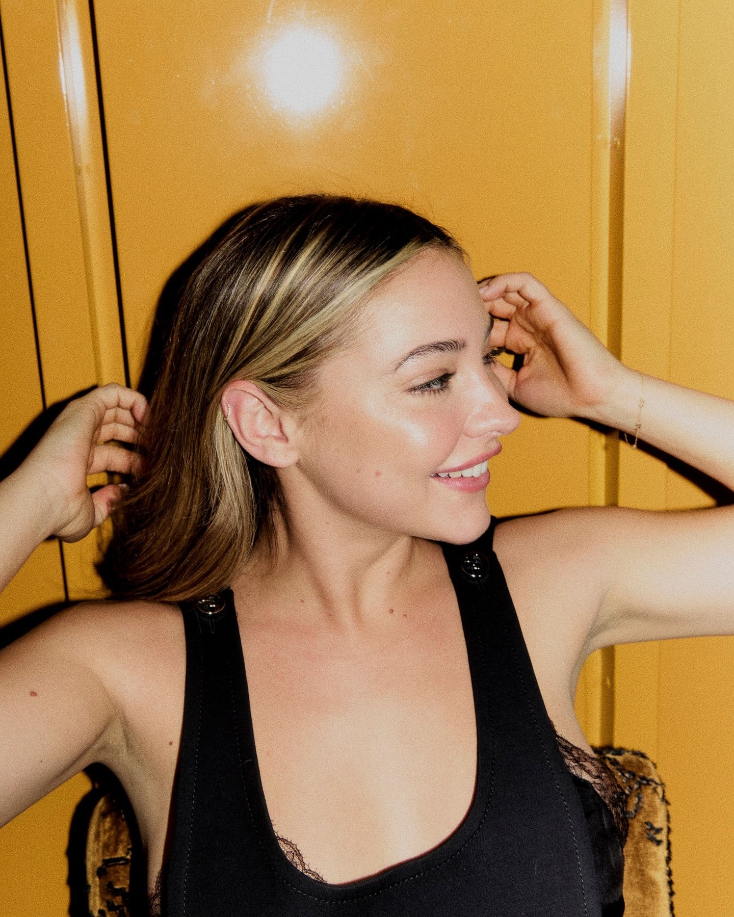 Smiling woman in a black tank top with blonde highlights against a yellow background. Shoulder-length hair frames her face in a close-up profile.