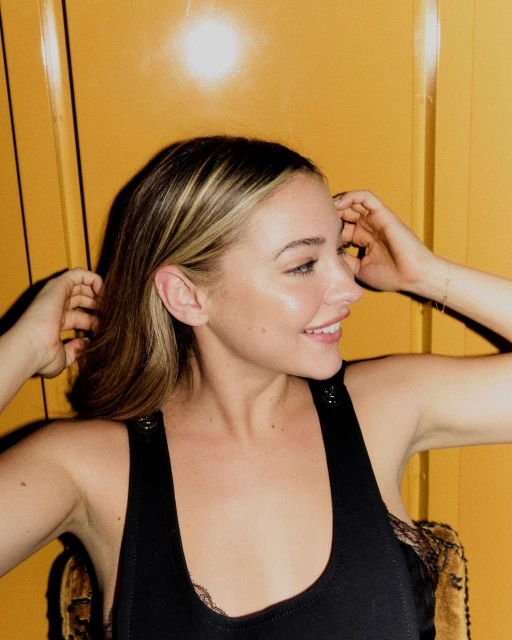 Smiling woman in a black tank top with blonde highlights against a yellow background. Shoulder-length hair frames her face in a close-up profile.