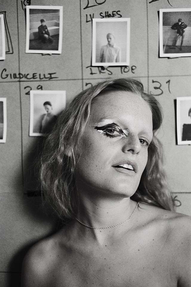 Black and white portrait of an adult female with dramatic eye makeup, posed in front of a wall of fashion reference photographs.