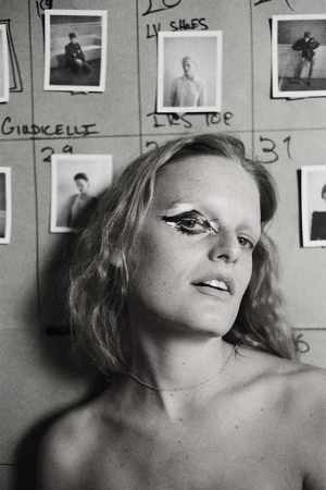 Black and white portrait of an adult female with dramatic eye makeup, posed in front of a wall of fashion reference photographs.