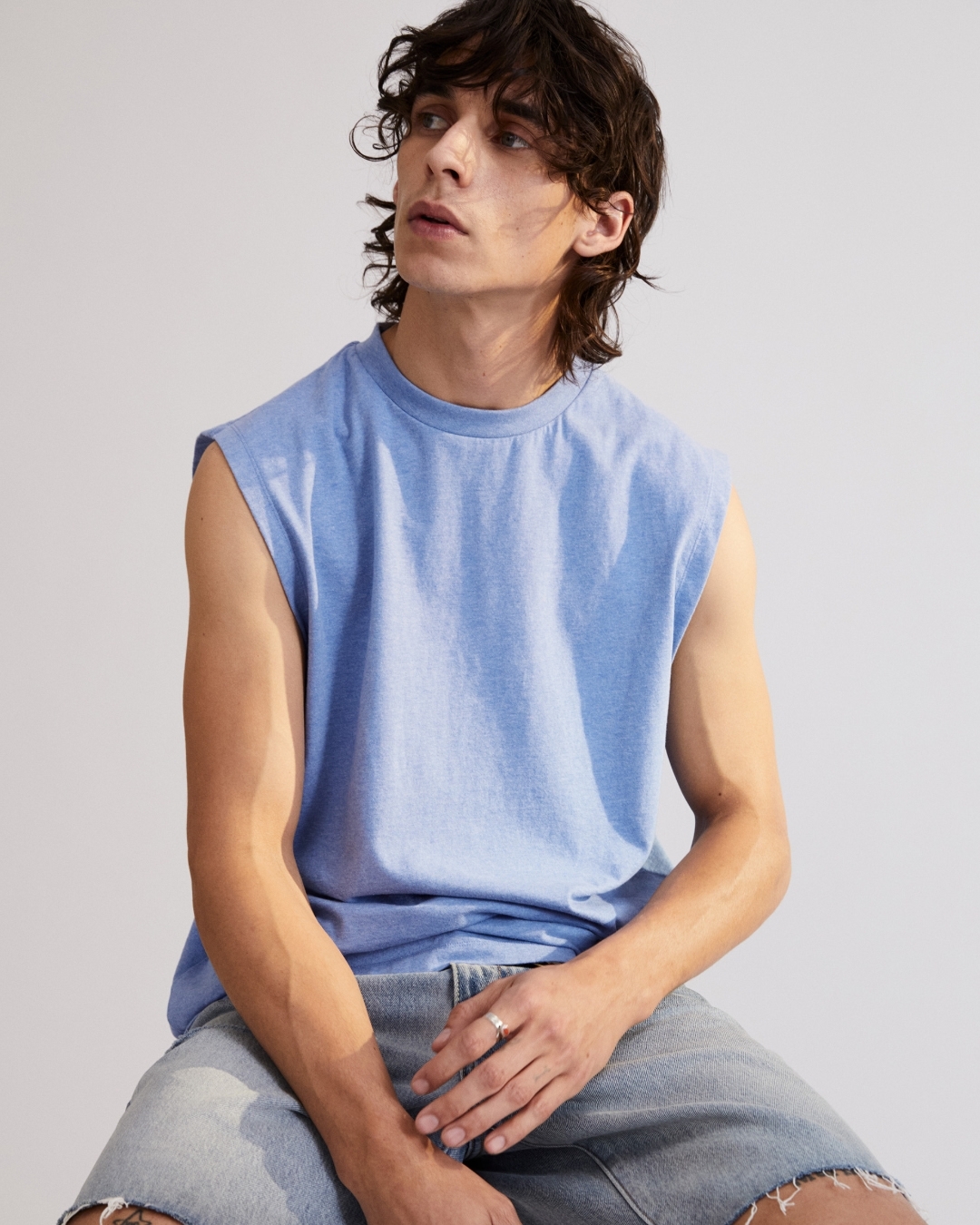 Young adult male with wavy brunette hair poses in a sleeveless blue shirt and ripped denim shorts in a studio setting.