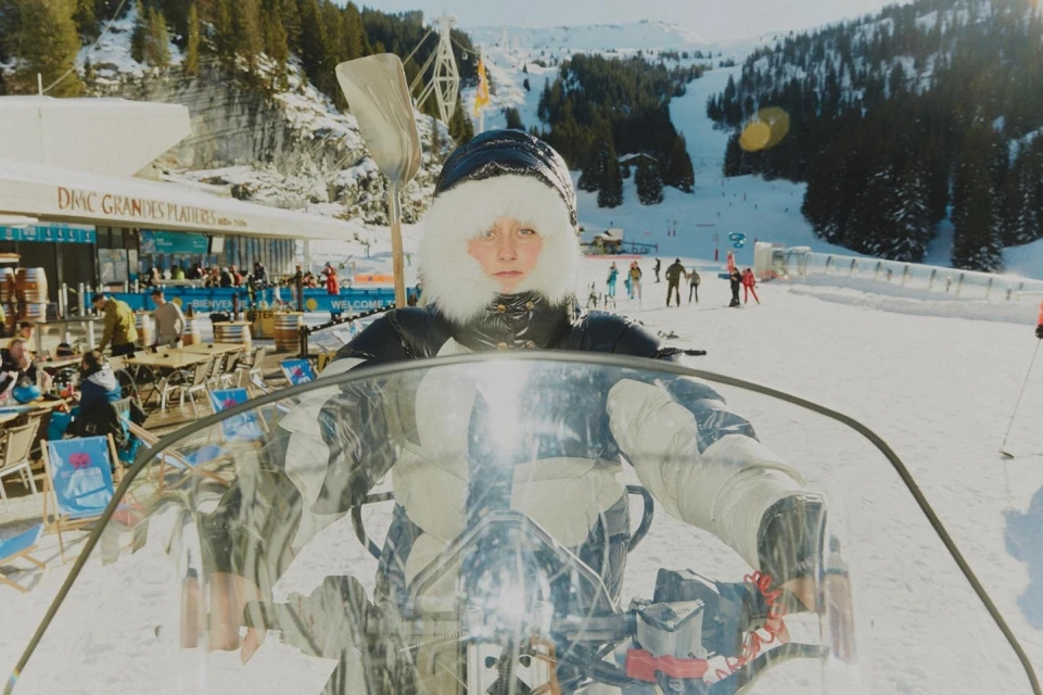 Woman in snow gear on a snowmobile at a ski resort, featuring a mountain backdrop, skiers, and architecture on a sunny day.