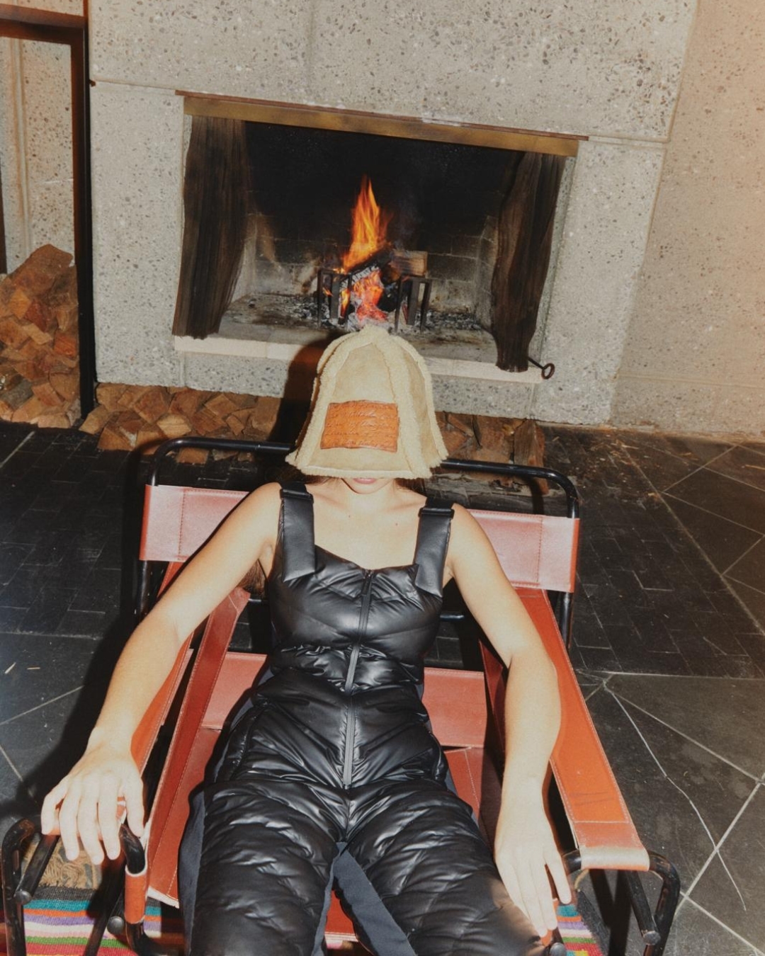 A person wearing a black jumpsuit and unique head covering sits in a chair in front of a stone fireplace with a fire burning.