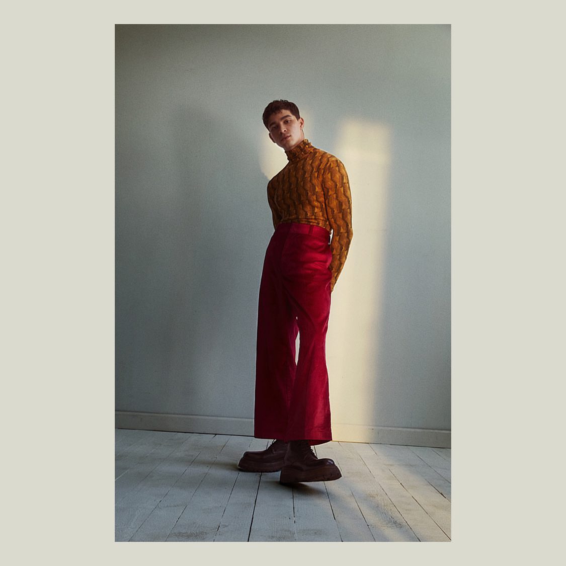 Adult male in patterned turtleneck and red pants stands in a brightly lit interior, posing for a fashion shot with neutral background.