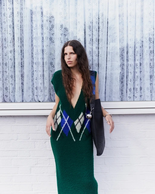 Adult female in a green, deep-V sleeveless argyle dress standing against a white brick wall near a window with lace curtains.
