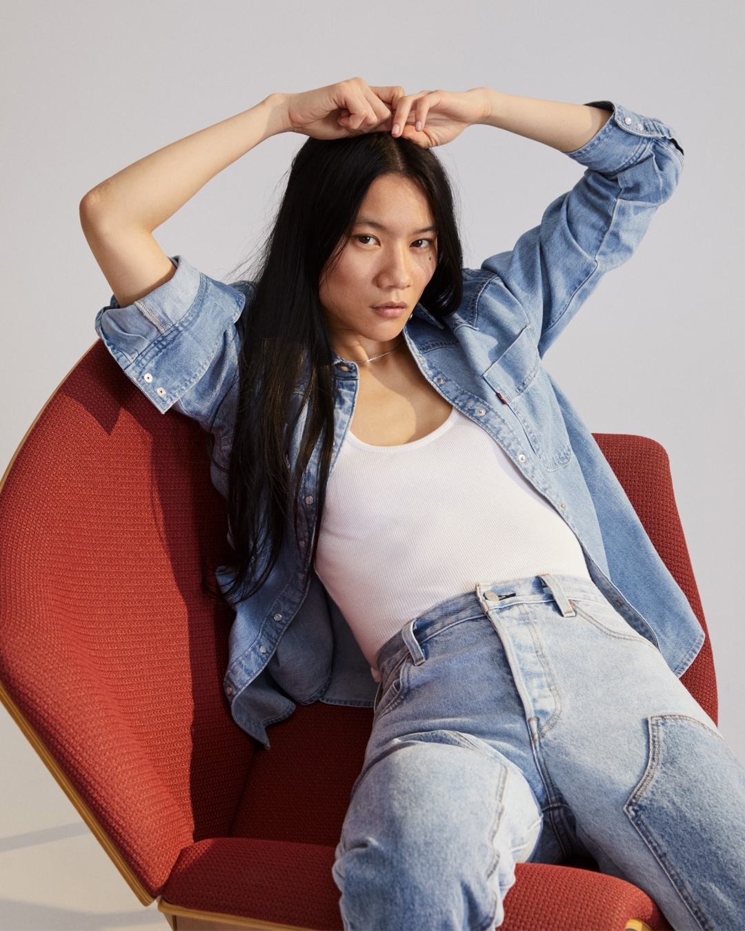 Portrait of an Asian woman in a denim jacket and jeans sitting in a red chair, posing with her hands above her head in a studio.