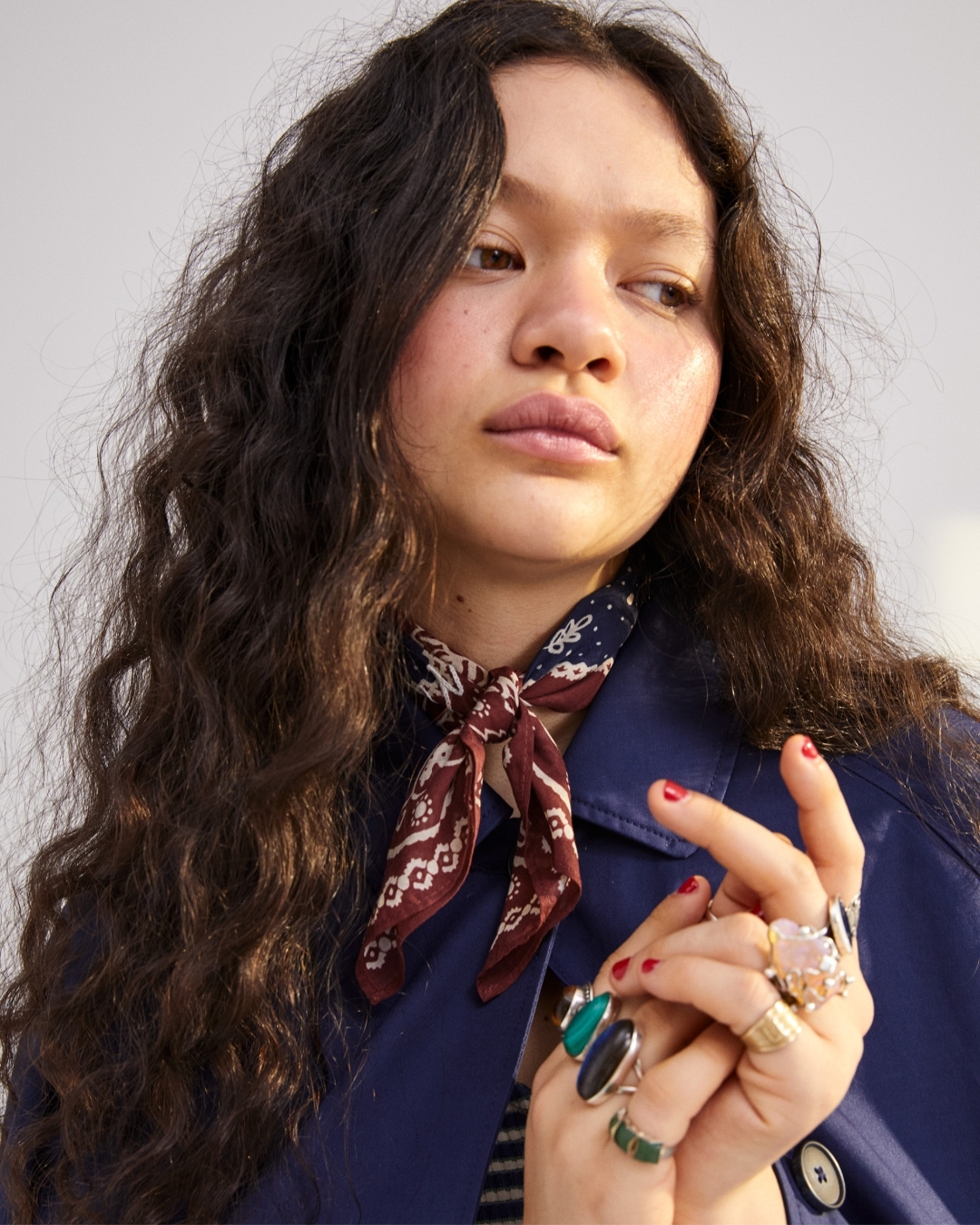 Portrait of an adult woman with dark curly hair wearing a blue jacket and printed neck scarf adorned with rings on her fingers.