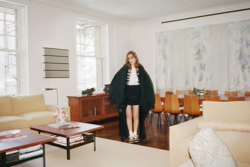 Young adult poses in a fashion editorial shoot inside a light-filled home with modern furniture, wearing a black coat and miniskirt.