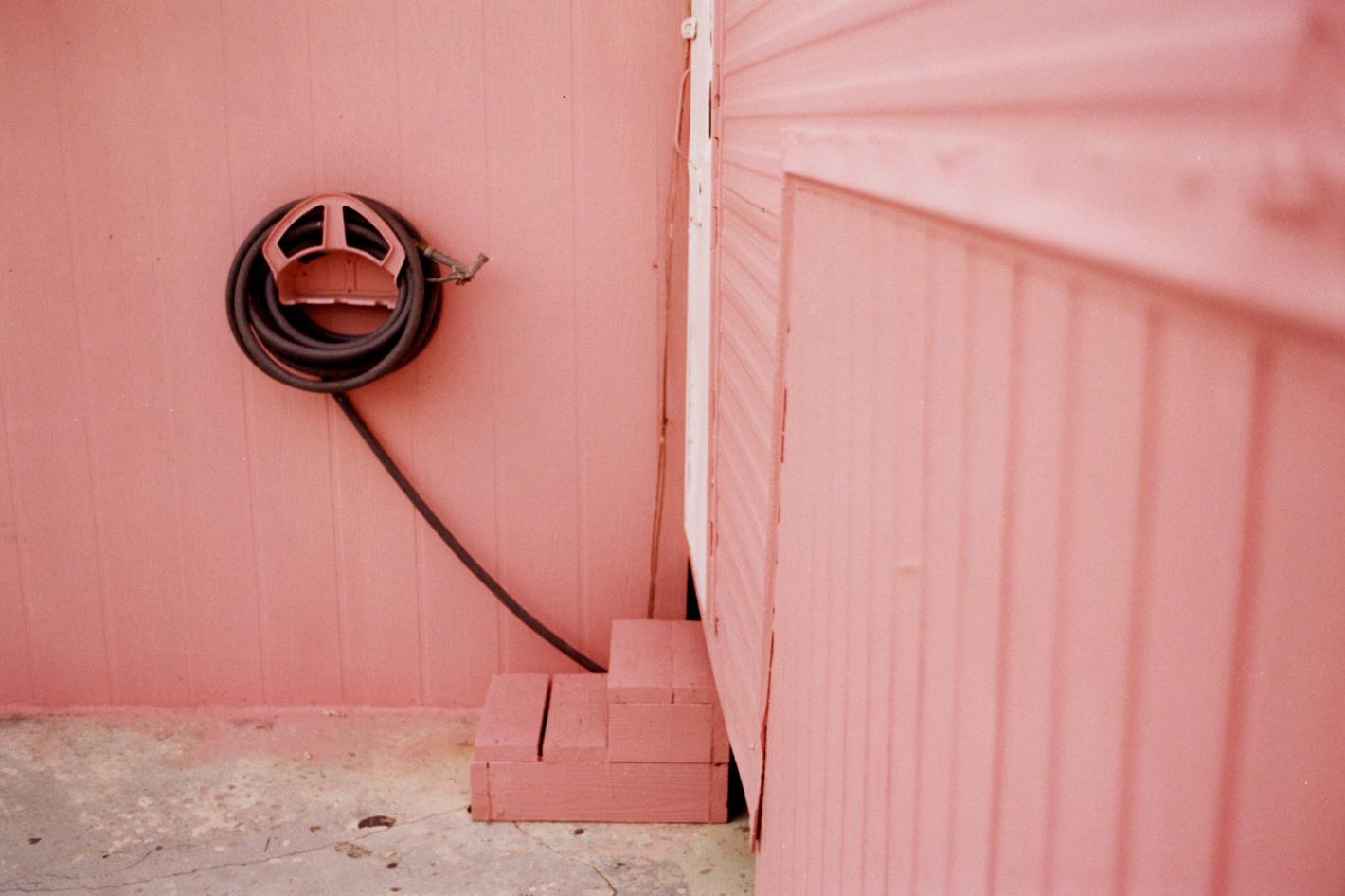 Exterior of a building featuring a pink wall, a neatly coiled black hose mounted on a bracket, and small wooden steps.