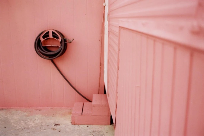 Exterior of a building featuring a pink wall, a neatly coiled black hose mounted on a bracket, and small wooden steps.