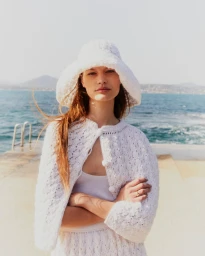 A woman in a white knitted bucket hat and white cardigan standing by the ocean under clear sky.