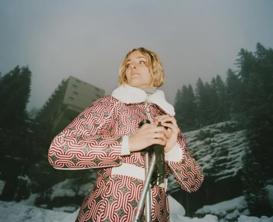 Woman in patterned red and white ski suit holding ski poles against a snowy mountain and overcast sky.