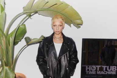 Model with bleached short hair and silver chain necklaces wearing a black leather jacket in front of a large palm leaf.
