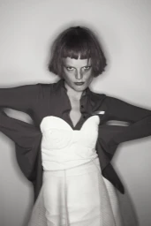 Portrait of a woman with a bob haircut wearing a collared top and layered skirt against a plain studio background.