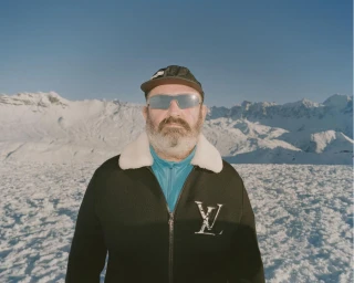 Man wearing sunglasses, black zip jacket, and cap against a snowy mountain landscape under a blue sky.