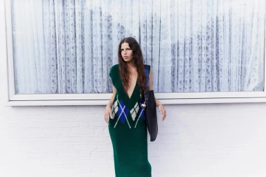 Woman with long dark hair wearing a green argyle knit dress leaning against a window with white lace curtains