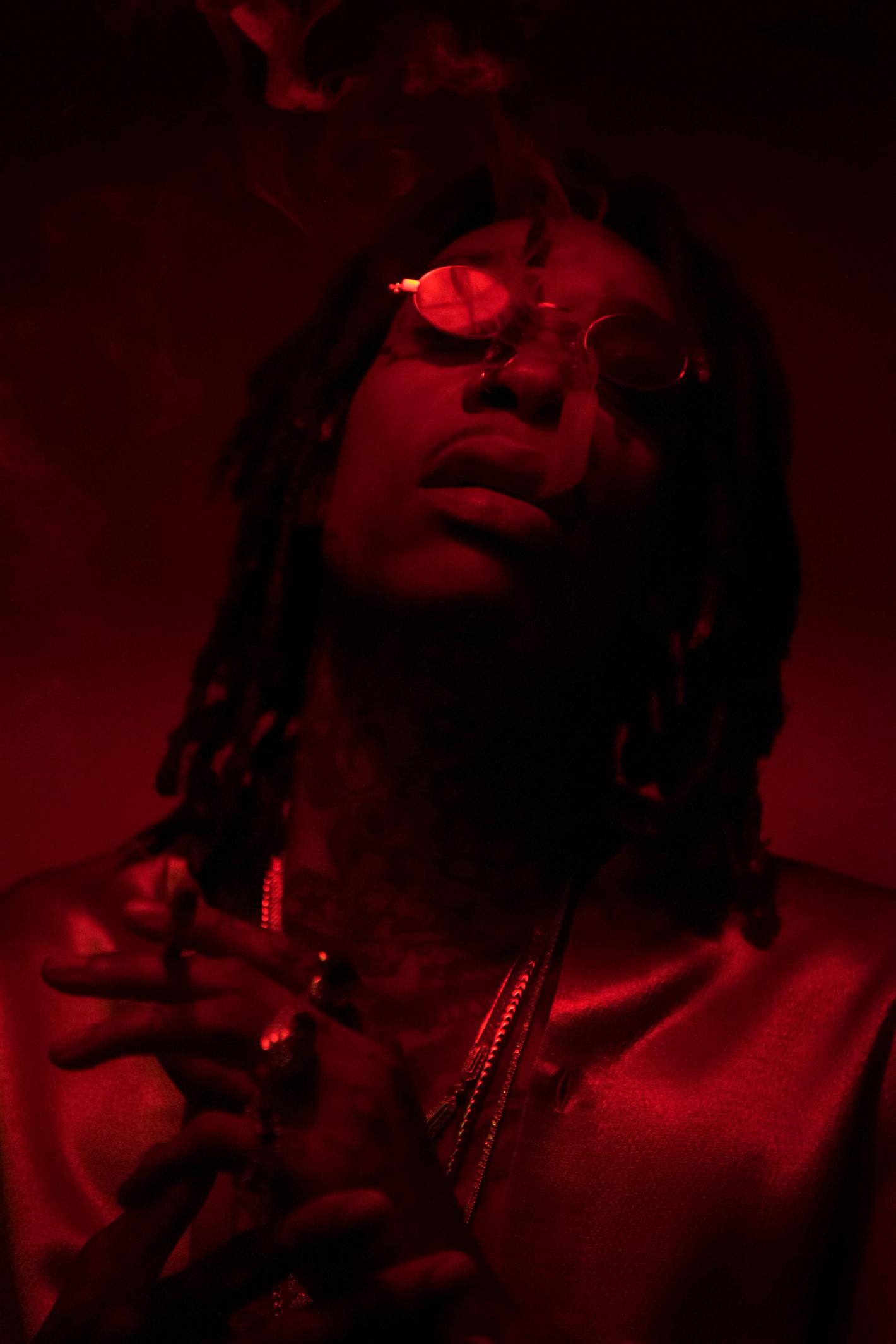 Low-angle portrait of an adult male with dreadlocks wearing round sunglasses and a gold chain, bathed in a dark red light with smoke.