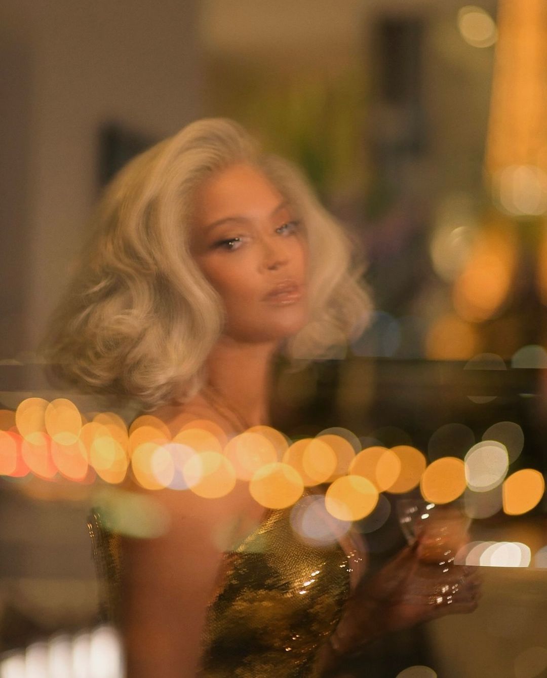 Elegant woman with platinum blonde hair in a sparkly gold dress, holding a drink, and surrounded by warm, bokeh lights indoors.