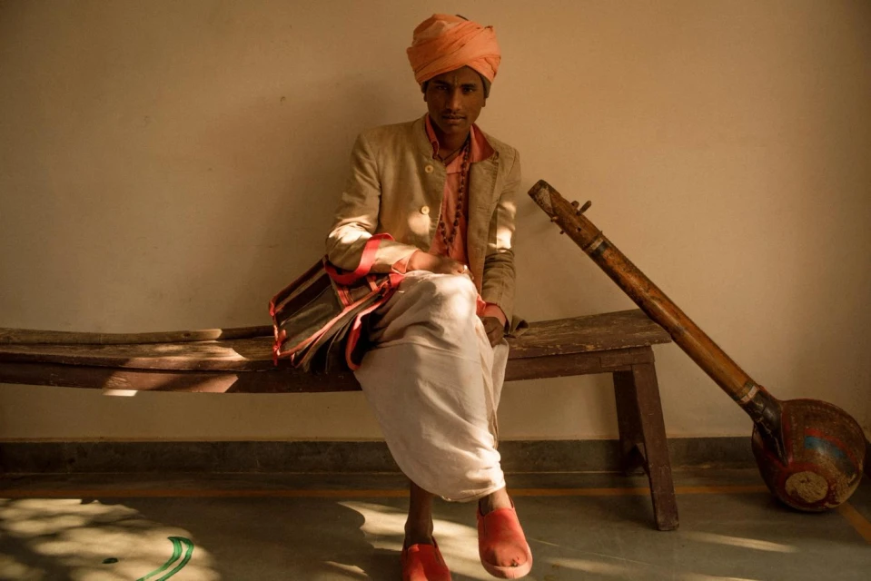Young man in traditional Indian clothing sits on a bench with a musical instrument in a softly lit indoor setting, creating a cultural portrait.