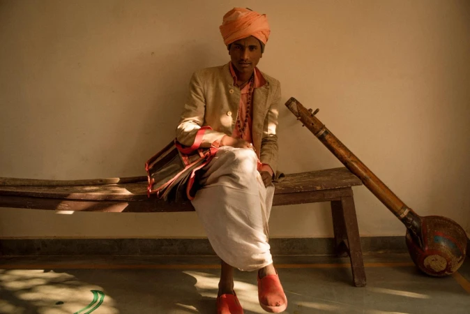 Young man in traditional Indian clothing sits on a bench with a musical instrument in a softly lit indoor setting, creating a cultural portrait.