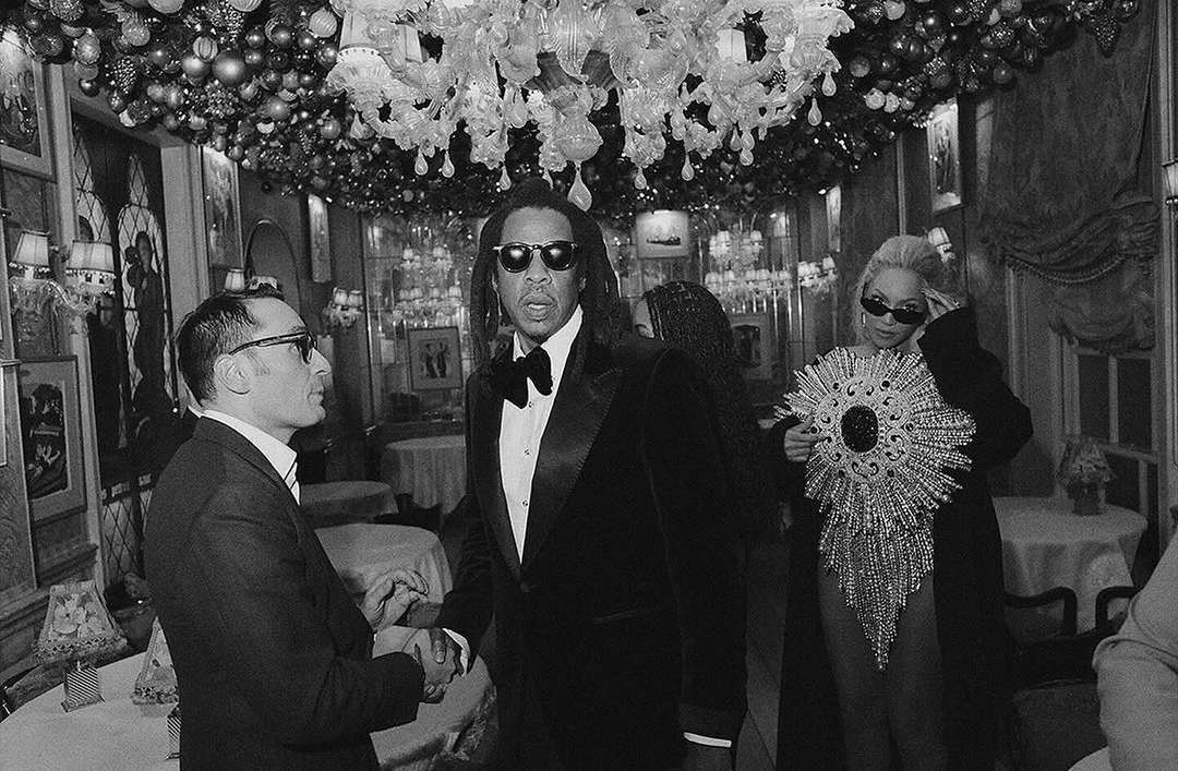 A black and white photo captures two men shaking hands in a restaurant with a chandelier, as two other adults stand nearby.