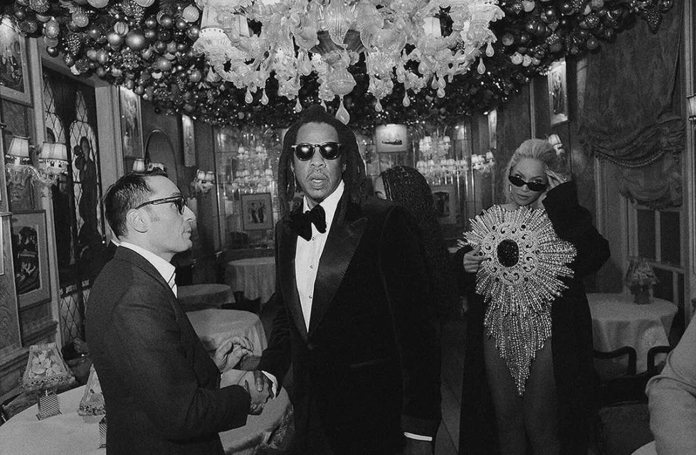 A black and white photo captures two men shaking hands in a restaurant with a chandelier, as two other adults stand nearby.