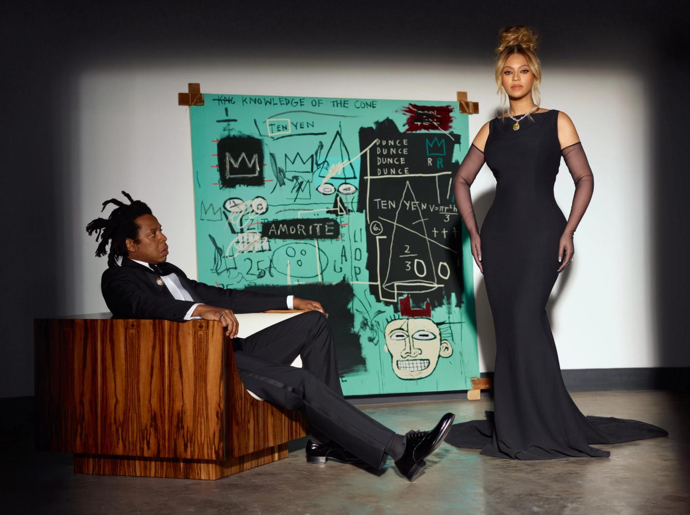 Beyonce in a long black dress stands next to Jay-Z sitting in a wood chair, posing in front of a colorful Jean-Michel Basquiat painting.