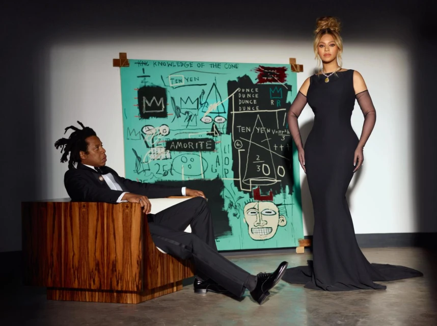 Beyonce in a long black dress stands next to Jay-Z sitting in a wood chair, posing in front of a colorful Jean-Michel Basquiat painting.