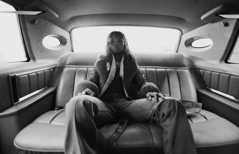 Monochrome portrait of an adult with dreadlocks, wearing a fur-lined coat, sitting in the back seat of a vintage car, interior shot.