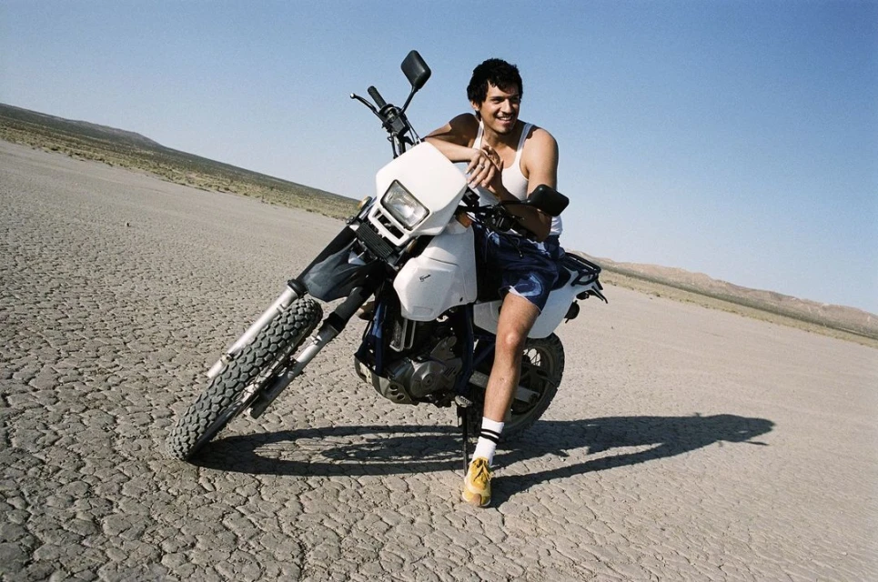 Man poses on dirt bike in desert landscape under clear sky, wearing white tank top, shorts, socks, and yellow shoes for an outdoor lifestyle shot.
