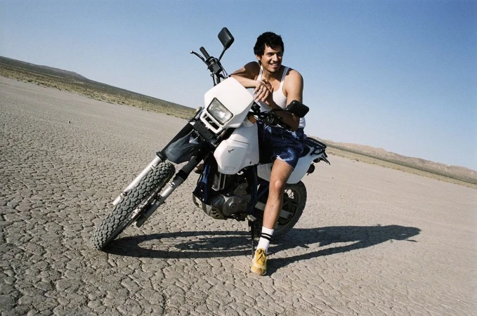 Man poses on dirt bike in desert landscape under clear sky, wearing white tank top, shorts, socks, and yellow shoes for an outdoor lifestyle shot.