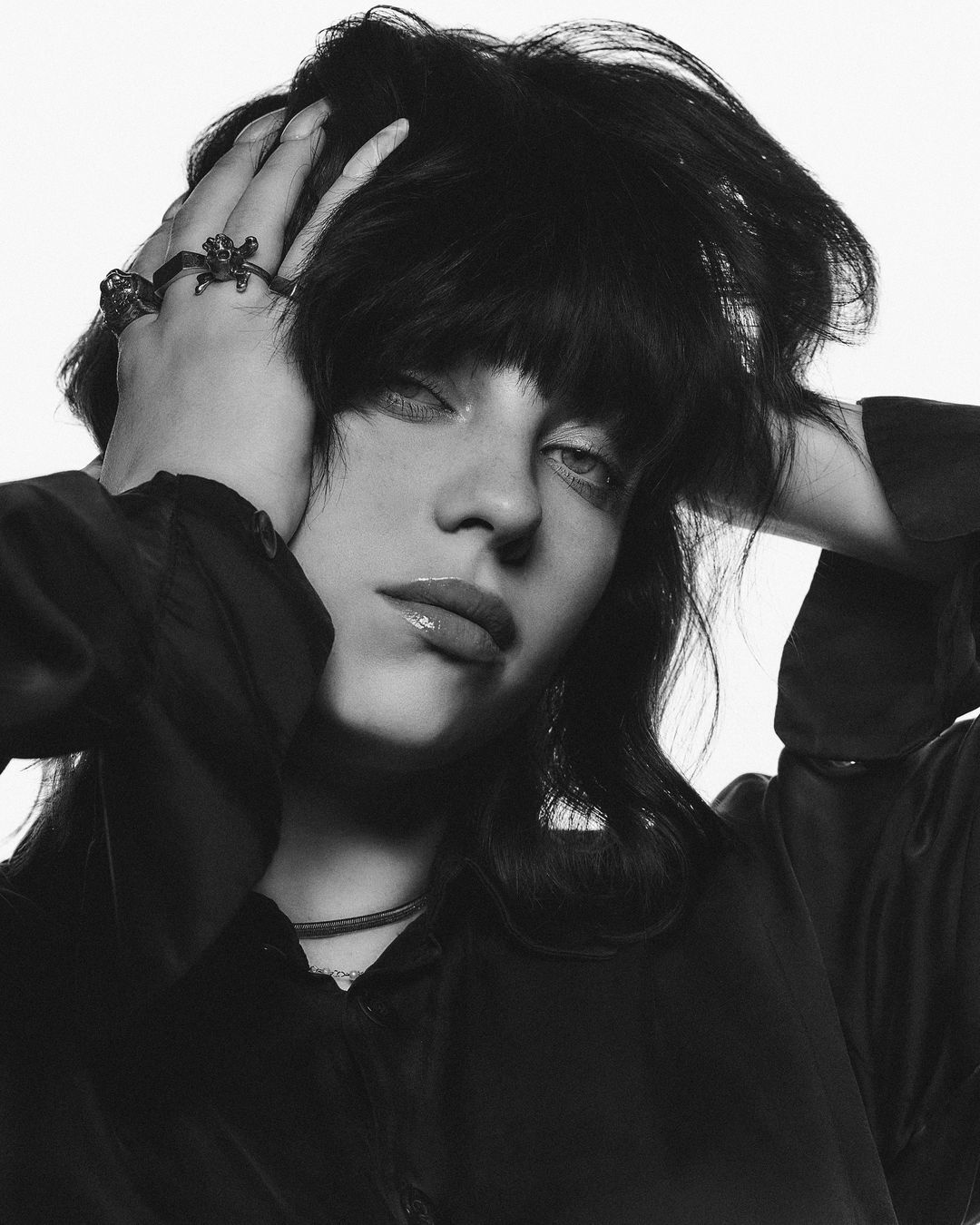 Striking black and white portrait of a young adult female with dark hair, hands on head, wearing a black shirt against a white backdrop.