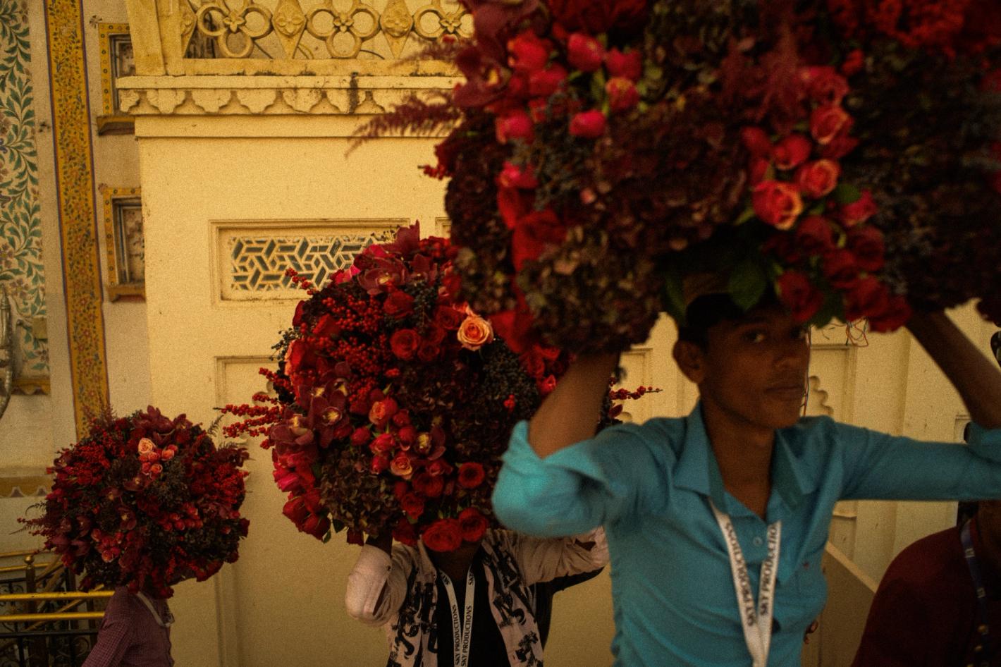 People carry large floral displays through an ornate building, with red roses and other flowers creating a vibrant, eye-catching spectacle.