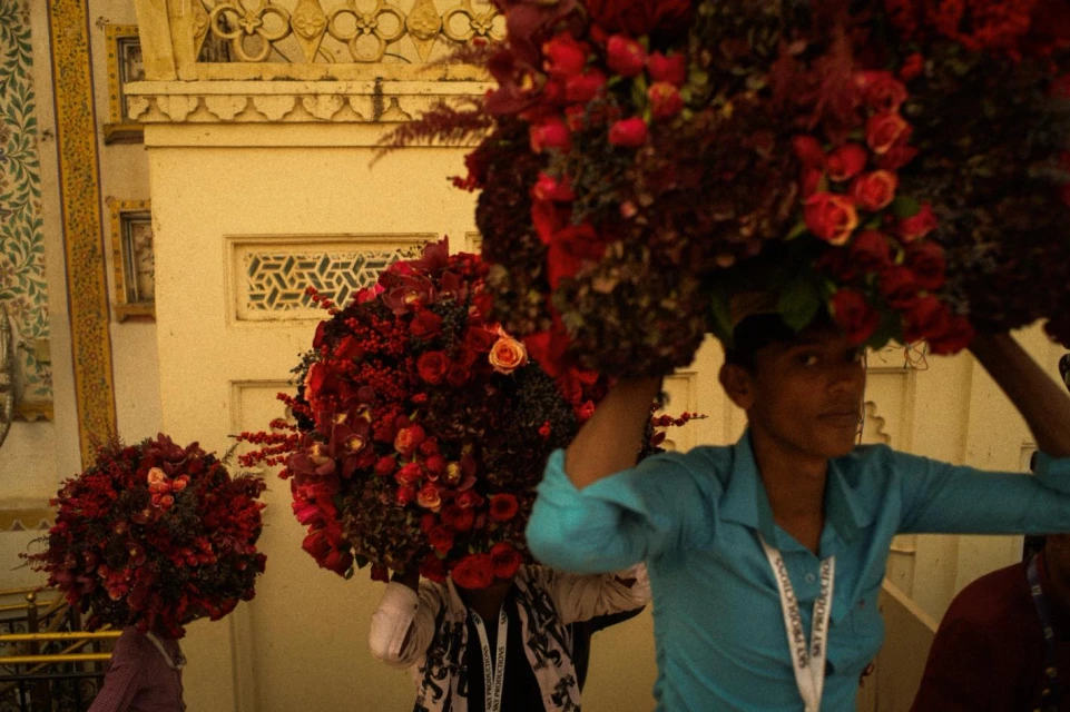 People carry large floral displays through an ornate building, with red roses and other flowers creating a vibrant, eye-catching spectacle.