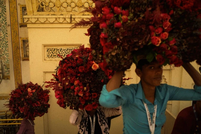 People carry large floral displays through an ornate building, with red roses and other flowers creating a vibrant, eye-catching spectacle.