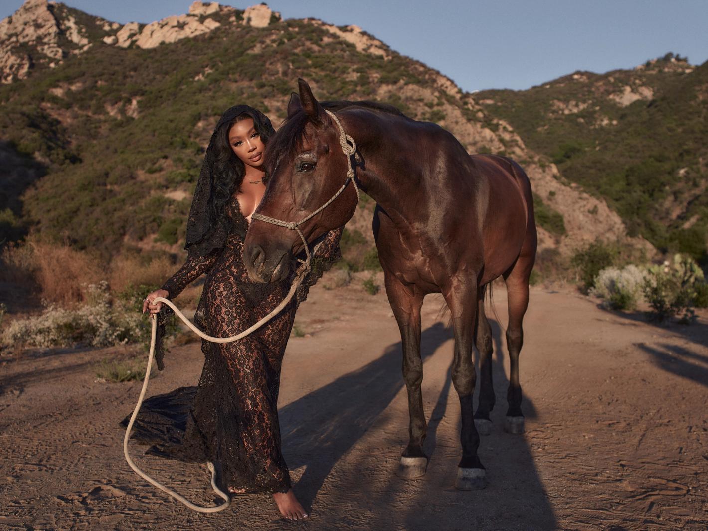 Woman in black lace gown poses with dark bay horse on a dirt road with mountain landscape in the background in warm light.