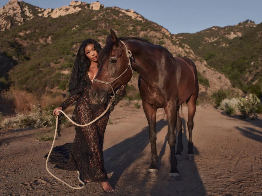 Woman in black lace gown poses with dark bay horse on a dirt road with mountain landscape in the background in warm light.