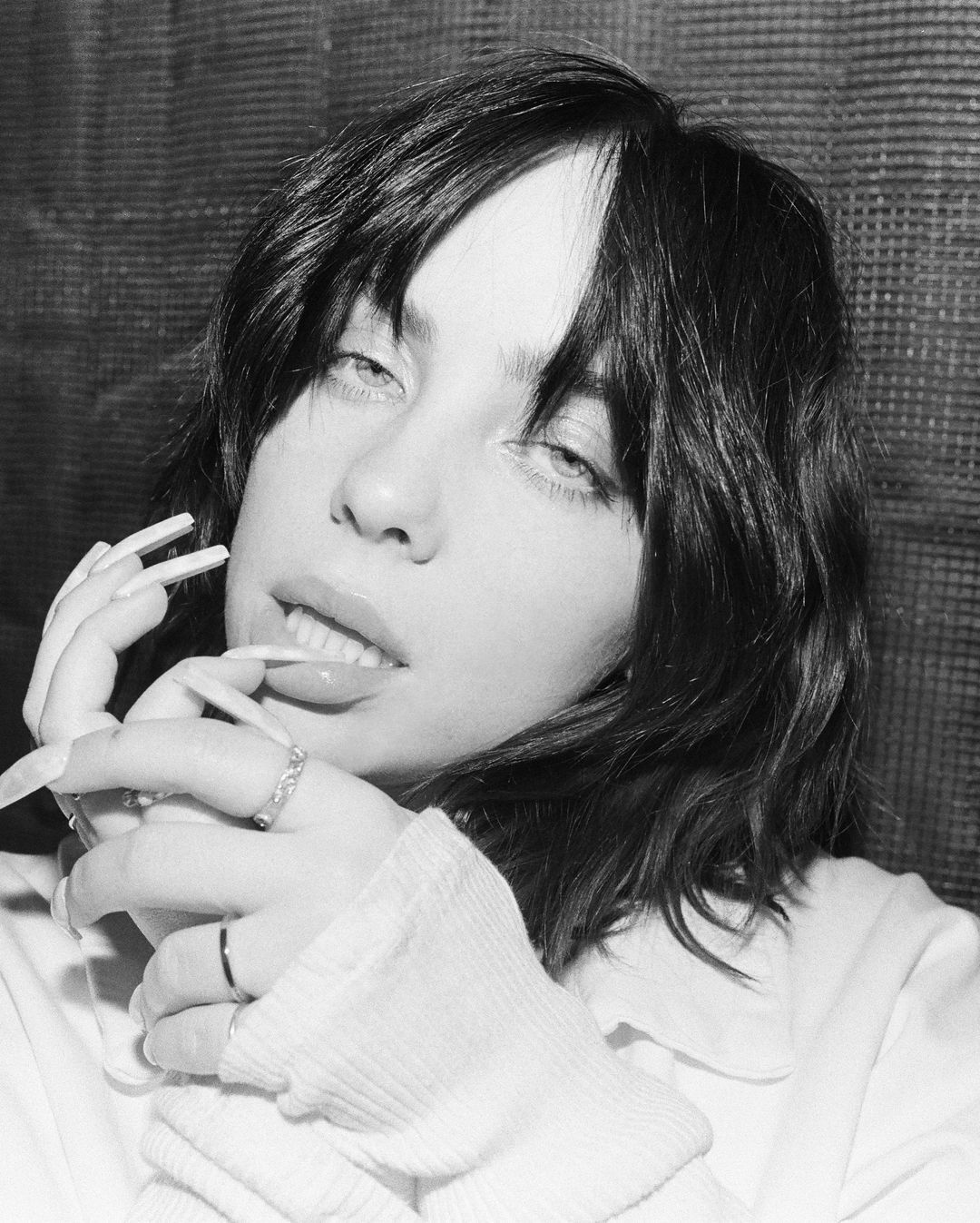Black and white portrait of a young woman with dark hair and light eyes, wearing a white shirt and rings, posing indoors.