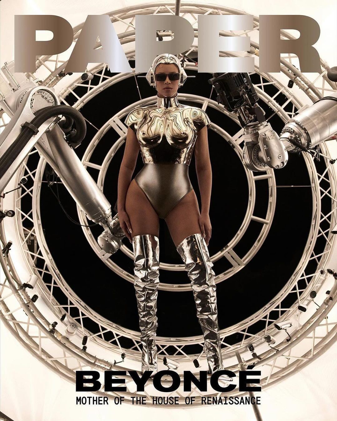 Beyoncé poses on the cover of Paper Magazine wearing a silver bodysuit and thigh-high boots against a backdrop of machinery.
