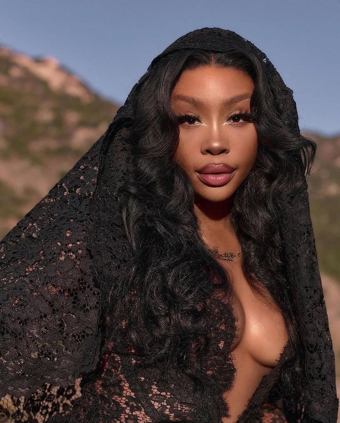 Close-up portrait of an adult woman wearing a black lace head covering against a background of blurred brown mountains and blue sky.