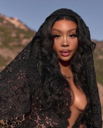 Close-up portrait of an adult woman wearing a black lace head covering against a background of blurred brown mountains and blue sky.