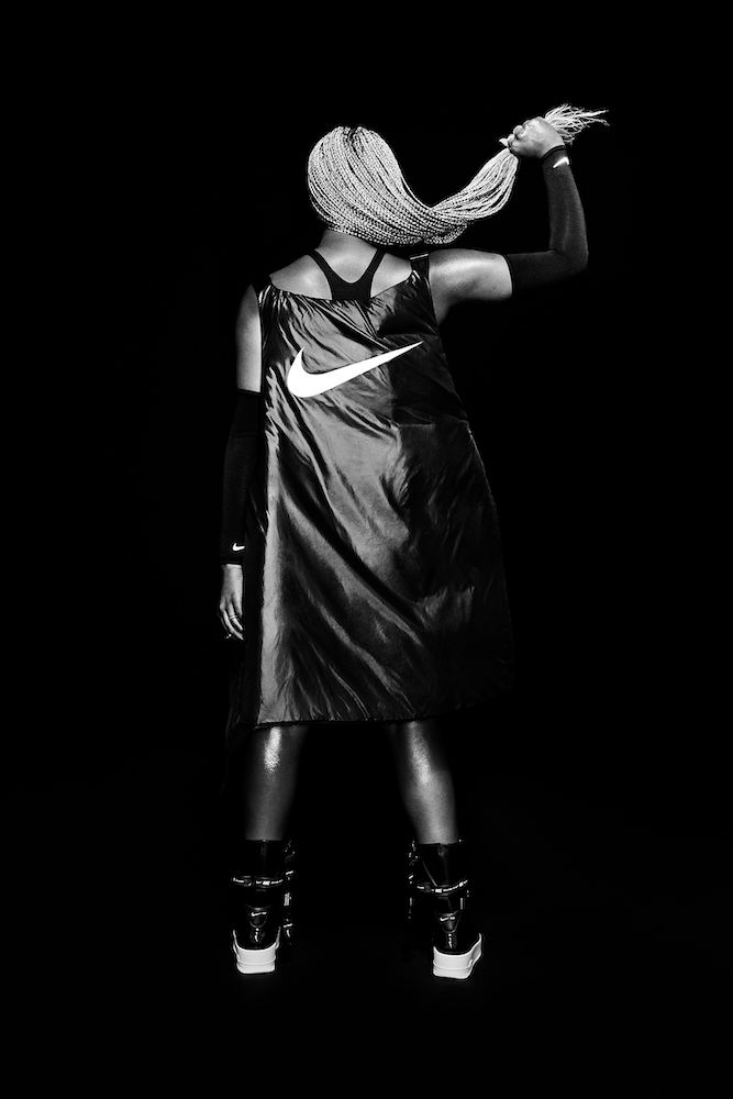 Back view of a woman with blonde braids holding her hair, wearing a black Nike jacket and shiny black boots against a dark background.