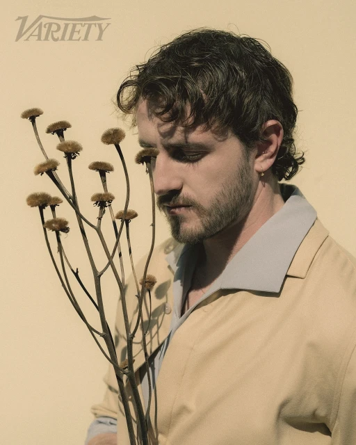 Portrait of fair-skinned man with beard holding dried floral arrangement against pale yellow background. He is wearing a tan jacket.