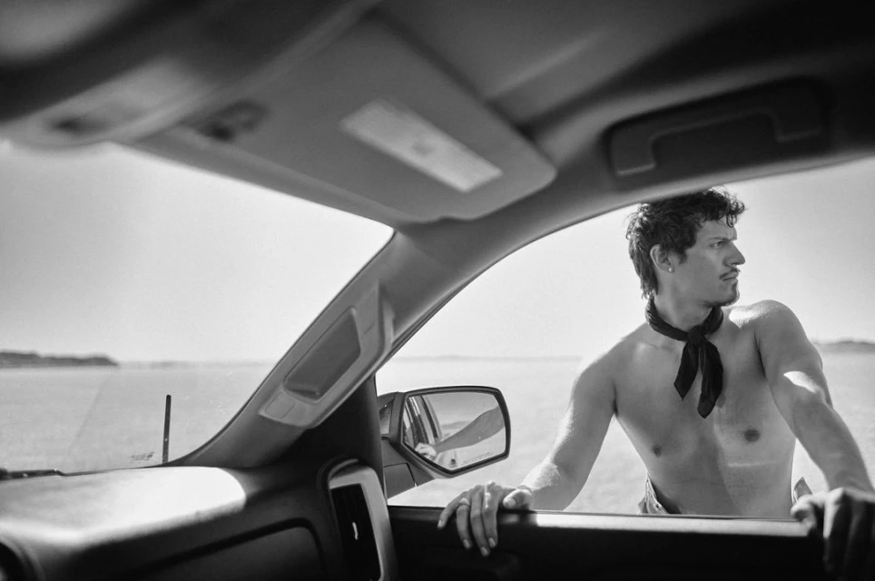 Black and white shot from inside a truck with an adult male wearing a bandana looking out over a sunlit flat landscape.