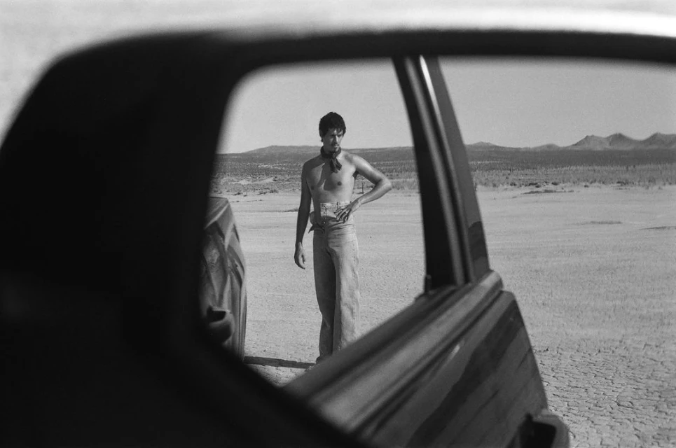 Black and white shot through car window of shirtless man standing in desert landscape with distant hills and cracked earth.