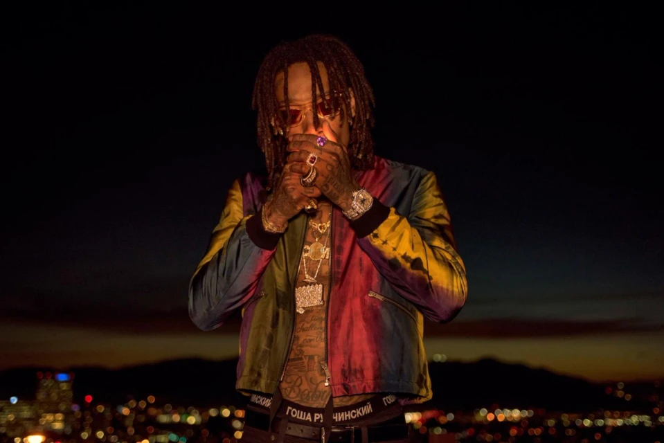 Man with dreadlocks in colorful jacket smoking in outdoor urban environment at night, city lights in background.