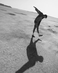 Monochrome image of a man leaping mid-air over a dry cracked desert terrain with a long shadow cast below by photographer Mason Poole.