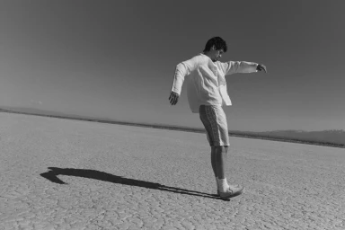 Monochrome photograph of a man balancing on one foot on a vast dry cracked salt flat under a clear sky.