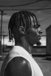 Black man in profile wearing a white sleeveless t-shirt and silver chain necklace against an outdoor backdrop