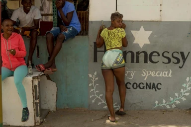A woman in a yellow lace top and patterned shorts standing by a painted wall with text, while seated people watch from a raised ledge nearby.